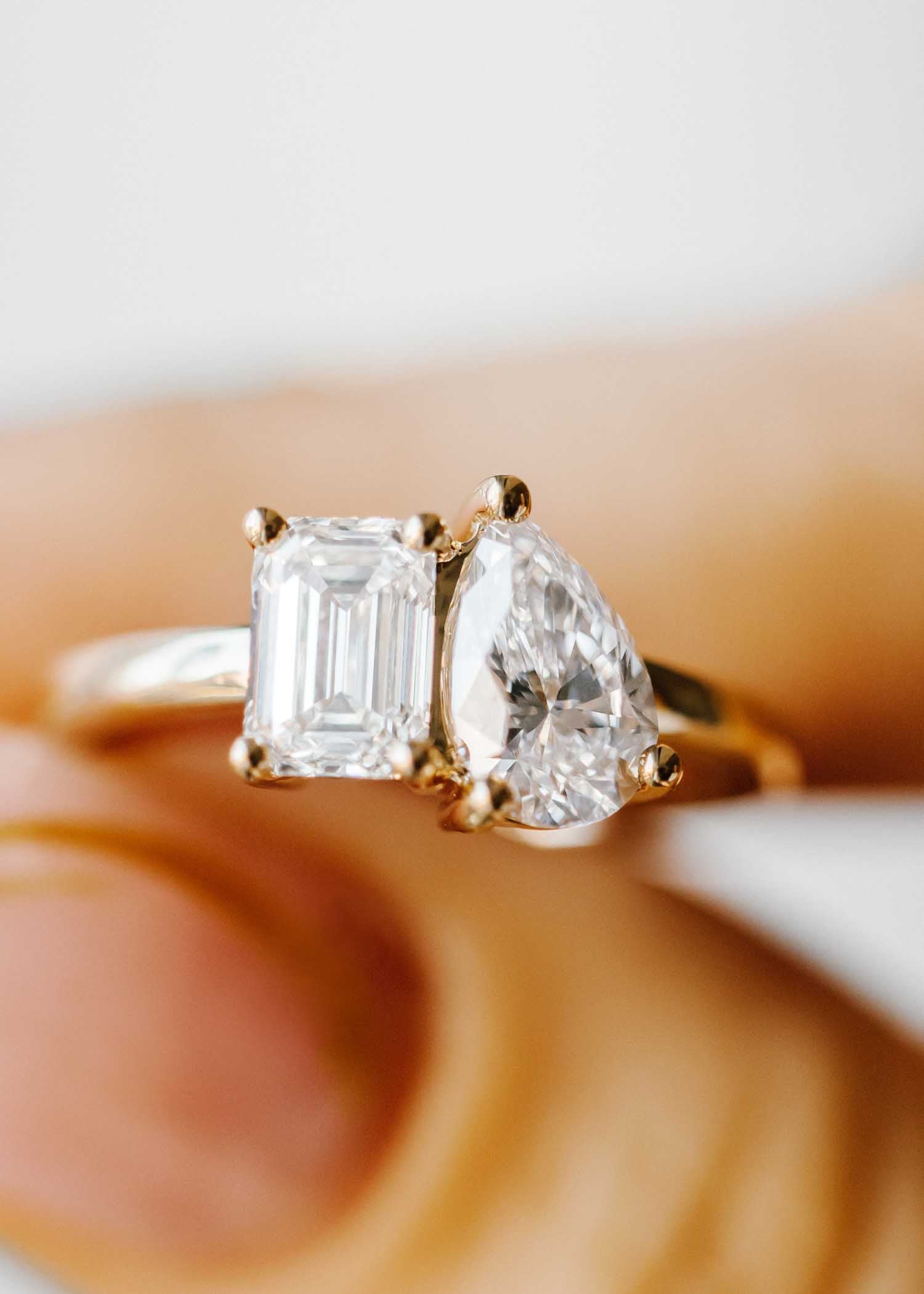 A close-up image of a custom-designed engagement ring featuring a stunning "Toi et Moi" design with two lab-grown diamonds. One diamond is emerald-cut, known for its elegant rectangular shape and step-cut facets. The other is pear-cut, celebrated for its teardrop shape and brilliance. Both diamonds are set in a yellow gold band, highlighting their clarity and brilliance. This two-stone ring, symbolizing "You and Me," is perfect for those seeking  sustainably sourced engagement rings.