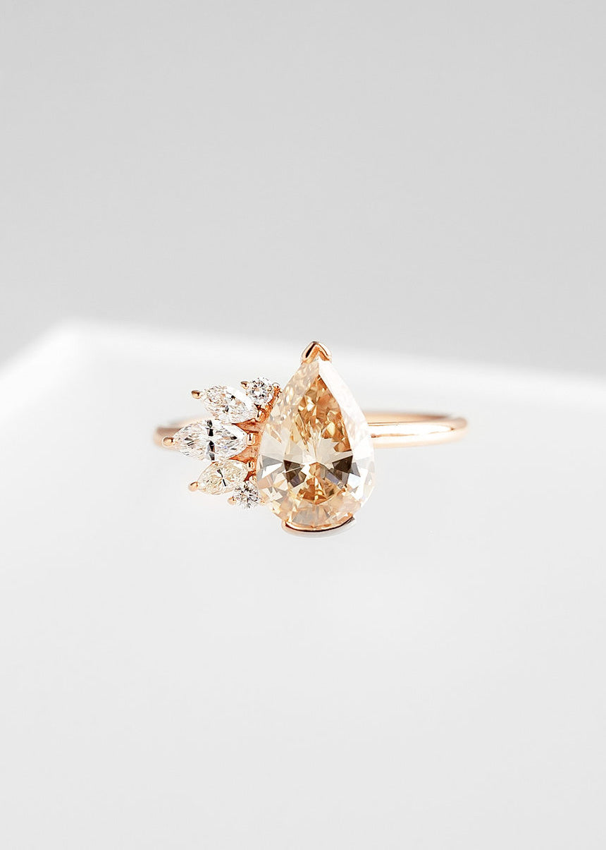 Exquisite rose gold engagement ring featuring a large pear-shaped champagne diamond at its center, complemented by a cluster of sparkling marquise and round diamonds on one side. The asymmetrical design adds a modern twist to the classic elegance, beautifully showcased against a soft grey background.