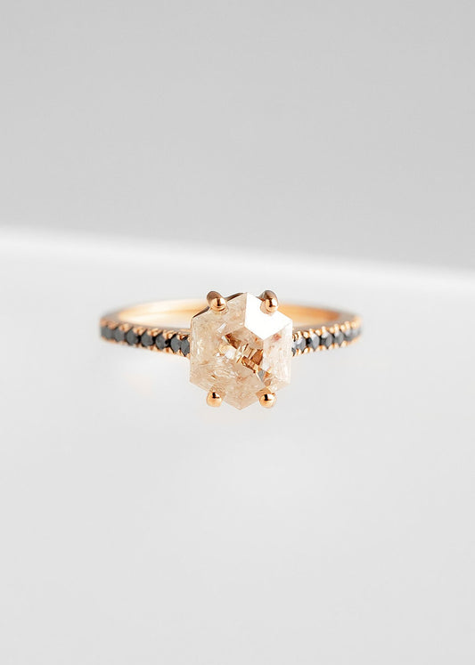 Elegant rose gold engagement ring featuring a prominent rose-colored salt and pepper diamond in a unique hexagonal cut, prong set to enhance its natural beauty. The band is adorned with a row of delicate black diamonds, adding a modern contrast to the overall design, all showcased against a soft grey background.