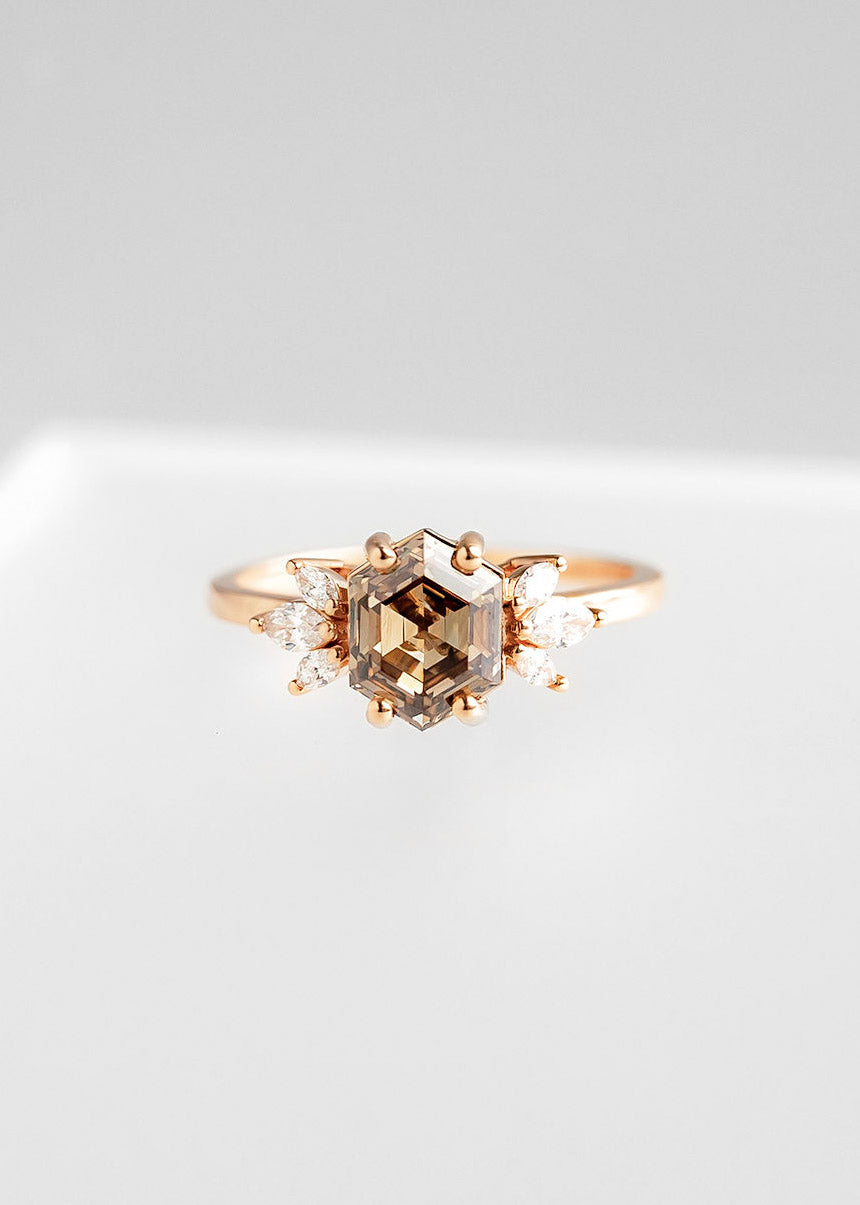 Stunning rose gold engagement ring featuring a central hexagon-cut champagne diamond, surrounded by symmetrical marquise-cut clear diamonds arranged in an elegant pattern. The design combines vintage charm with modern sophistication, beautifully displayed against a soft grey background to highlight its unique and timeless appeal.