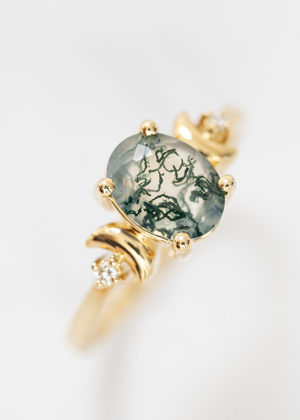 Elegant yellow gold engagement ring showcasing a large, oval-cut moss agate stone, set in prongs that highlight its translucent quality and vibrant green inclusions resembling moss. The design is enhanced by two crescent moons formed on each side of the ring, incorporating small round diamonds that add a touch of sparkle. The entire piece is presented against a crisp white background, emphasizing the ring's organic beauty and unique celestial motifs.