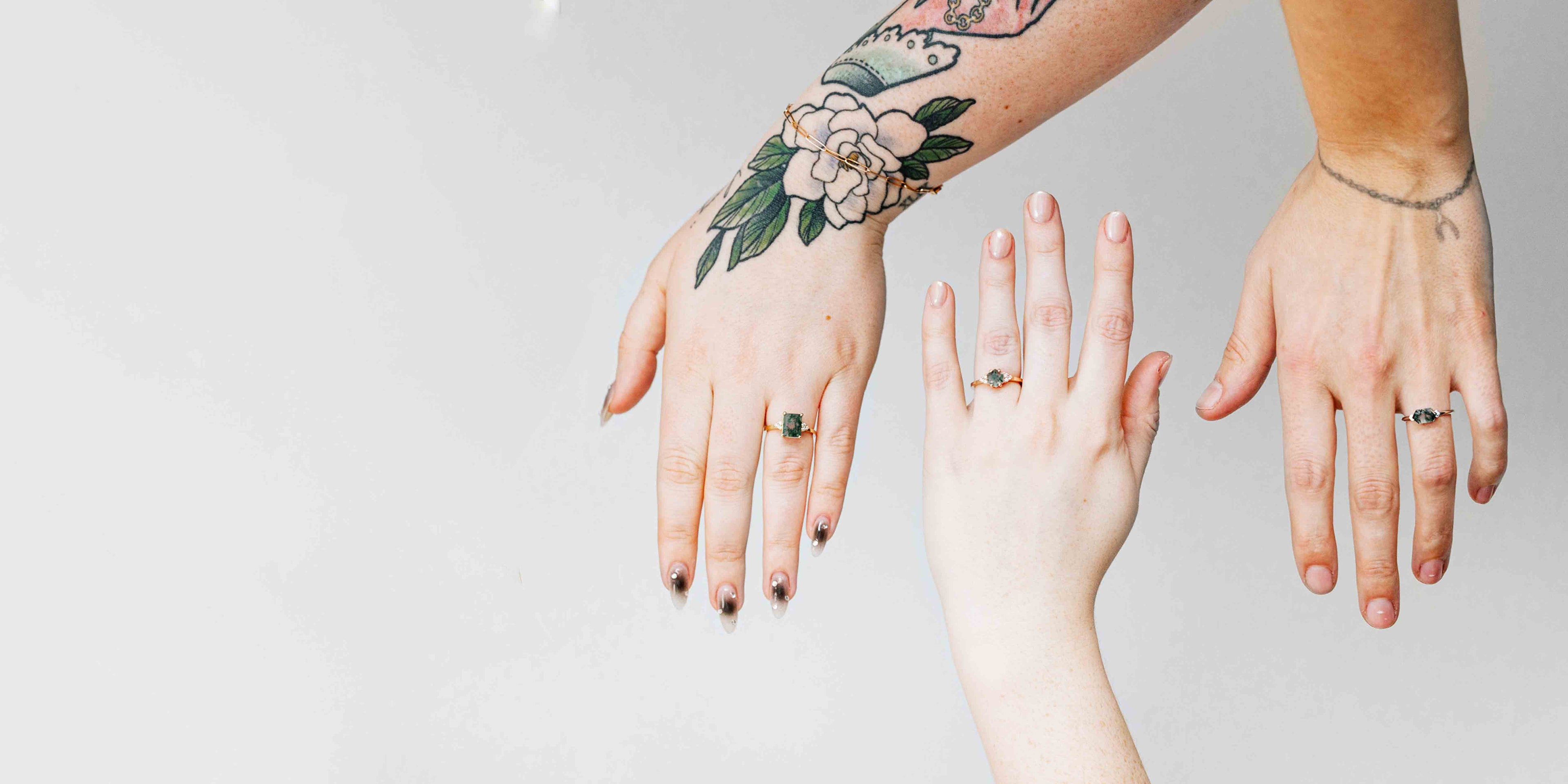 Three hands showcasing unique engagement rings with moss agate stones against a white background. The first hand features a tattooed floral design and displays a square moss agate in a gold setting. The second hand, with a rose gold band and small stones, extends from the right, and the third hand shows a east-west set moss agate ring, also with minimal tattoos.